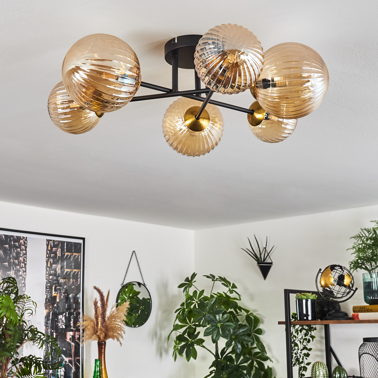 Eine moderne Deckenlampe mit sechs bernsteinfarbenen Glaskugeln hängt von einer weißen Decke. Der Raum hat Pflanzen, einen Spiegel und ein Bild.