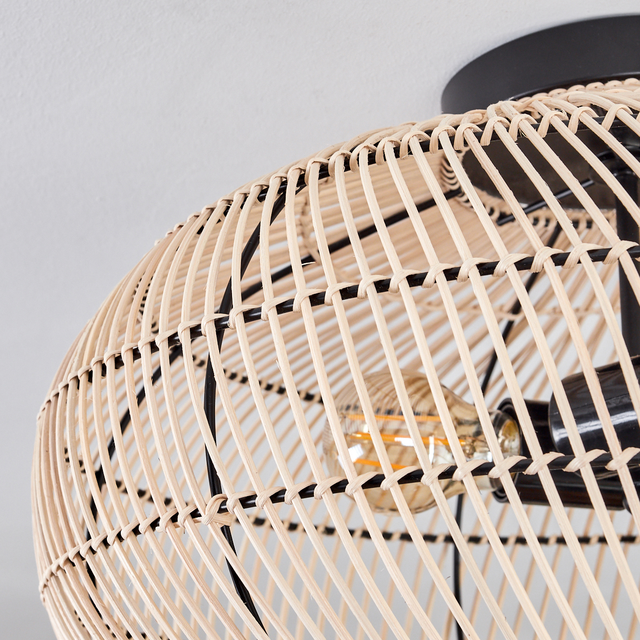 Nahaufnahme eines Lampenschirms aus geflochtenem Rattan mit schwarzem Metallrahmen. Im Inneren ist eine Glühbirne zu sehen. Der Hintergrund ist eine weiße Decke.