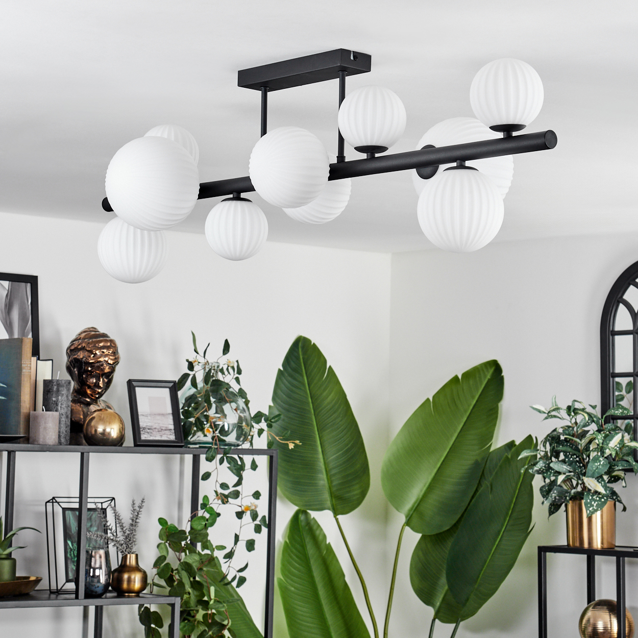 Ein modernes Zimmer mit einer schwarzen Deckenleuchte mit weißen Kugeln, großen grünen Pflanzen und Dekorationsartikeln auf Regalen.
