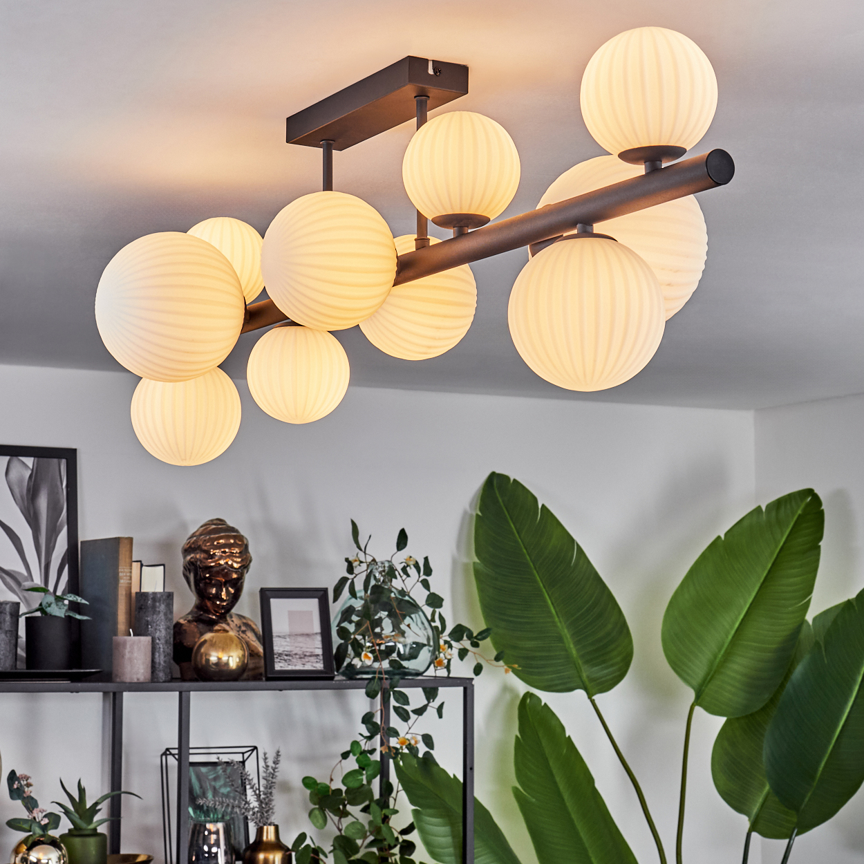 Eine moderne Deckenleuchte mit mehreren kugelförmigen Lampen. Darunter ein Regal mit Pflanzen und Dekorationsartikeln. Große grüne Blätter rechts.
