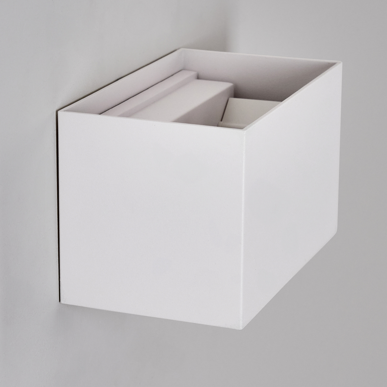 Eine weiße, rechteckige Box an einer grauen Wand. Im Inneren befinden sich weiße, rechteckige Objekte. Die Box hat eine offene Oberseite.