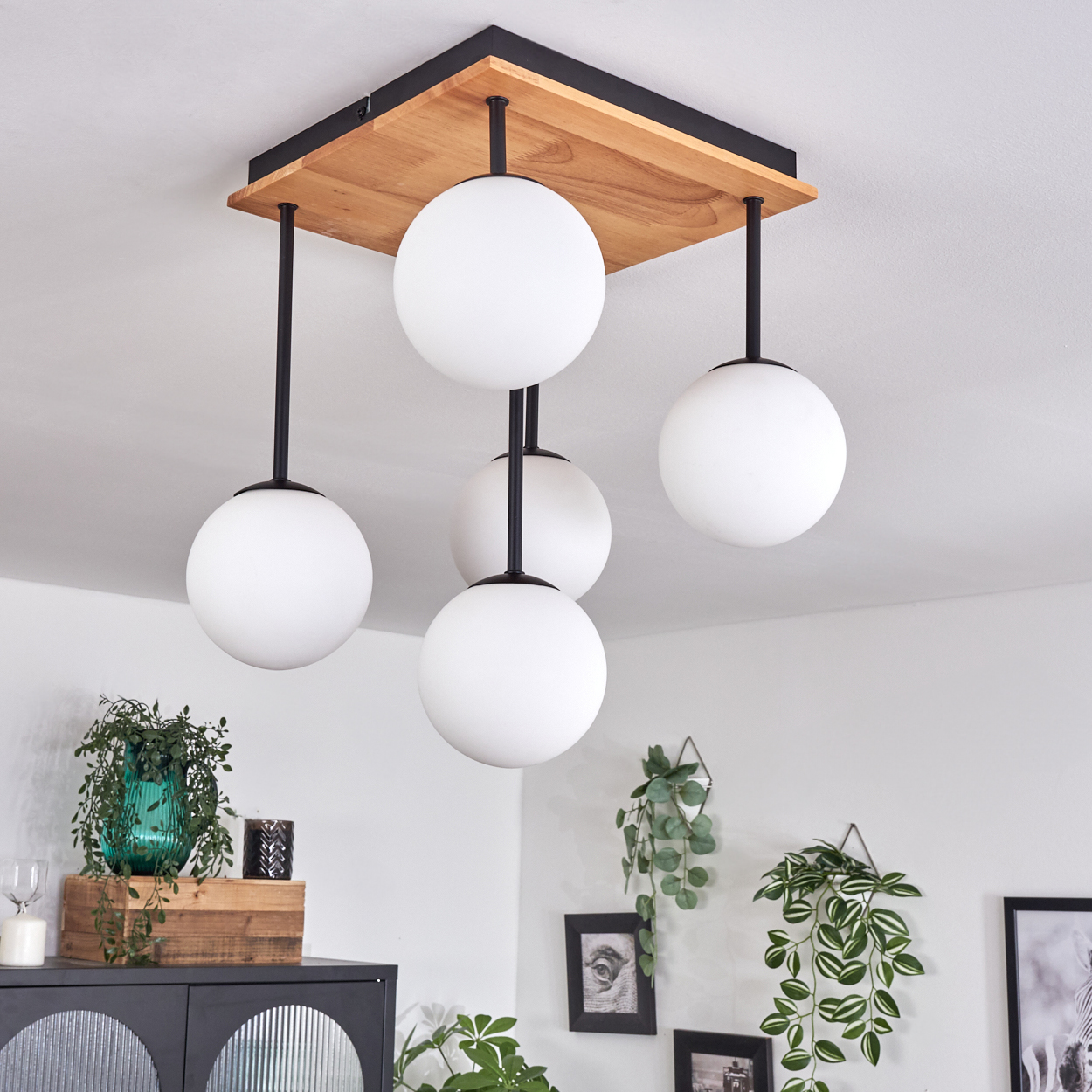 Moderne Deckenleuchte mit fünf weißen Kugelleuchten. Ein Holzpaneel trägt die Leuchten. Pflanzen und Möbel sind im Hintergrund.