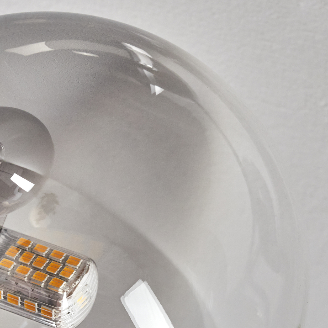 Nahaufnahme einer Glühbirne. Die Glühbirne ist klar und hat eine gebogene Form. Eine LED-Leuchte ist sichtbar. Der Hintergrund ist hellgrau.