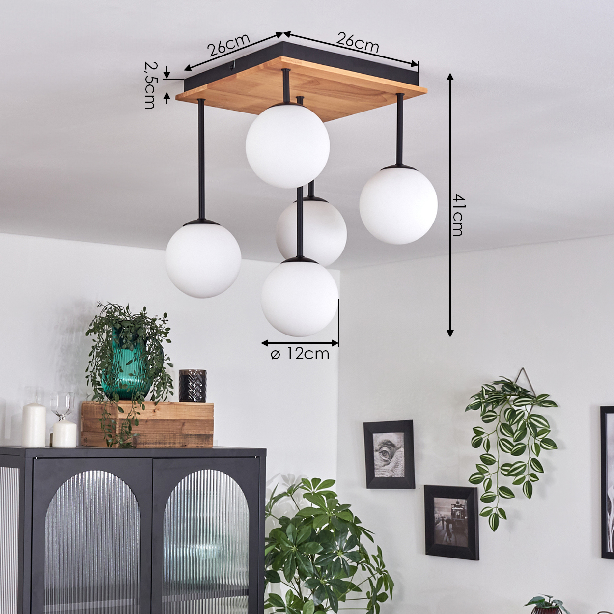 Eine Deckenlampe mit 4 weißen Kugelleuchten. Ein quadratischer Sockel aus Holz und Schwarz. Ein Schrank mit Deko und Pflanzen im Hintergrund.