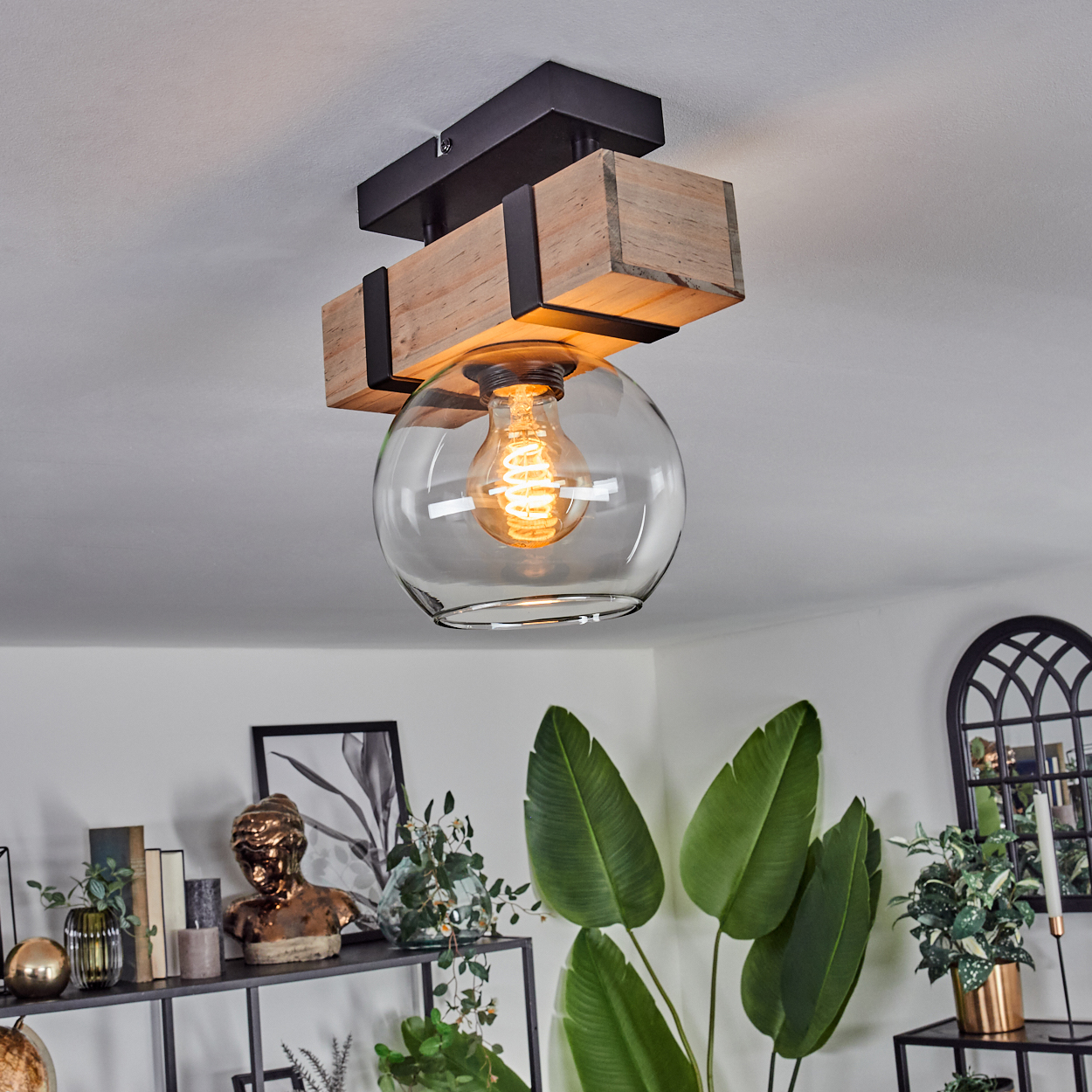 Eine Deckenlampe mit Holz- und Metallakzenten. Ein Glaskolben umschließt eine leuchtende Glühbirne. Dekorative Pflanzen und ein Regal mit Dekor im Hintergrund.