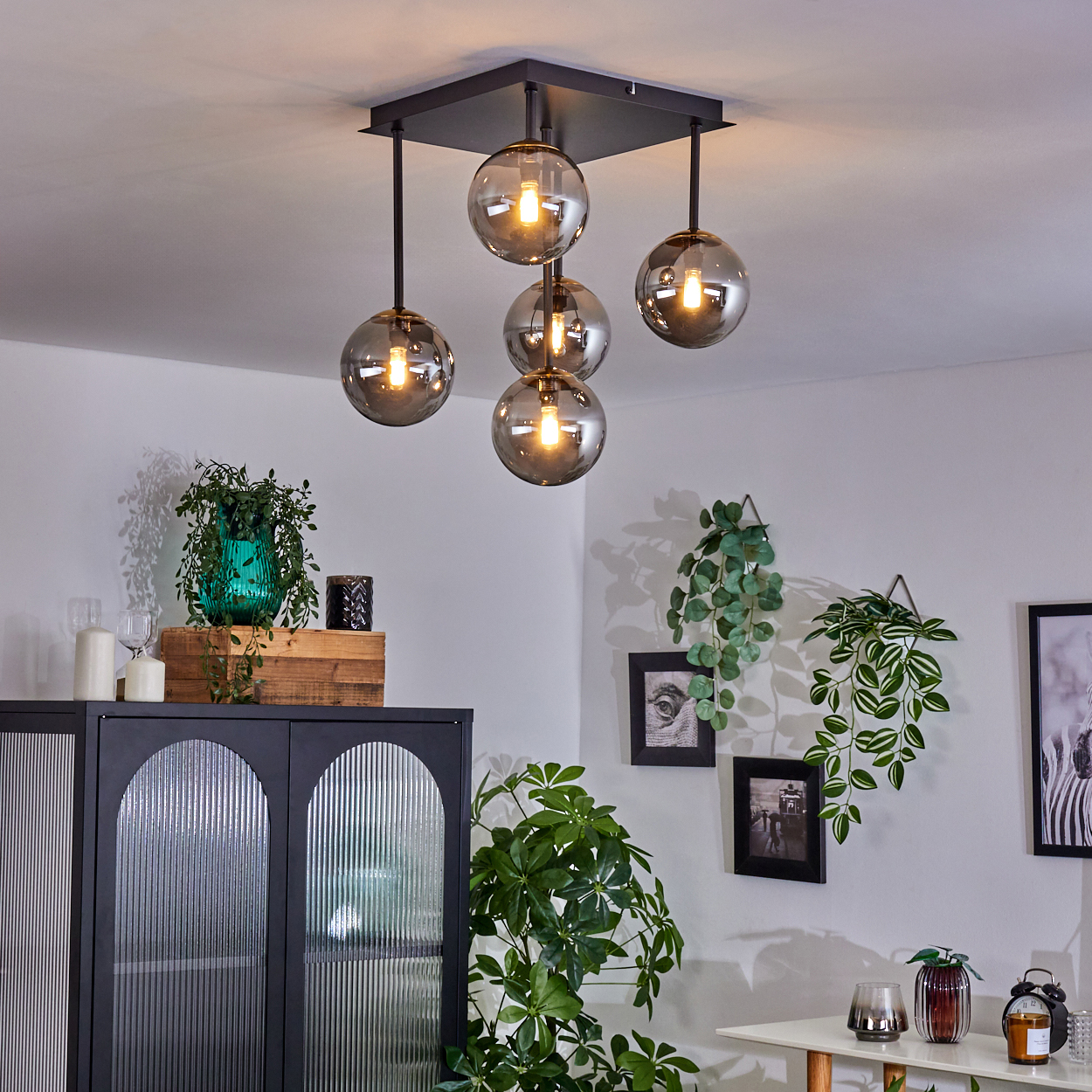 Modernes Wohnzimmer mit Deckenleuchte mit fünf Kugelleuchten. Pflanzen, Bilder und ein schwarzer Schrank sind ebenfalls sichtbar.