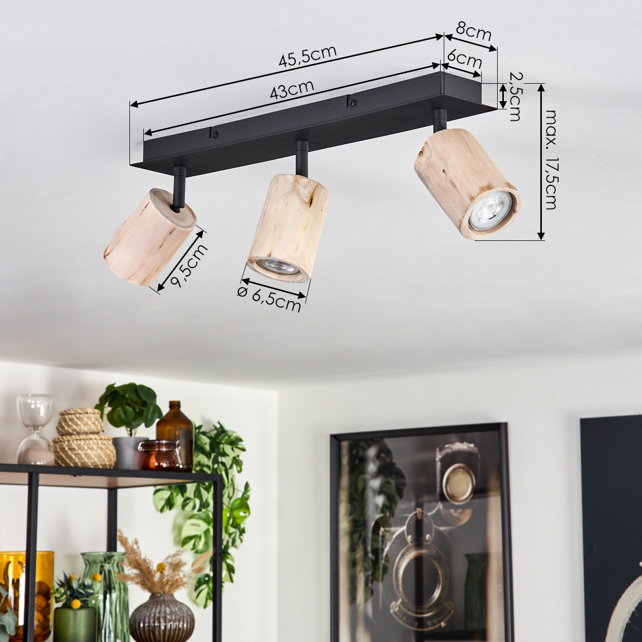 Eine Deckenleuchte mit drei Holzlampen. Abmessungen sind beschriftet. Hintergrund zeigt Deko mit Regalen und einem gerahmten Bild.
