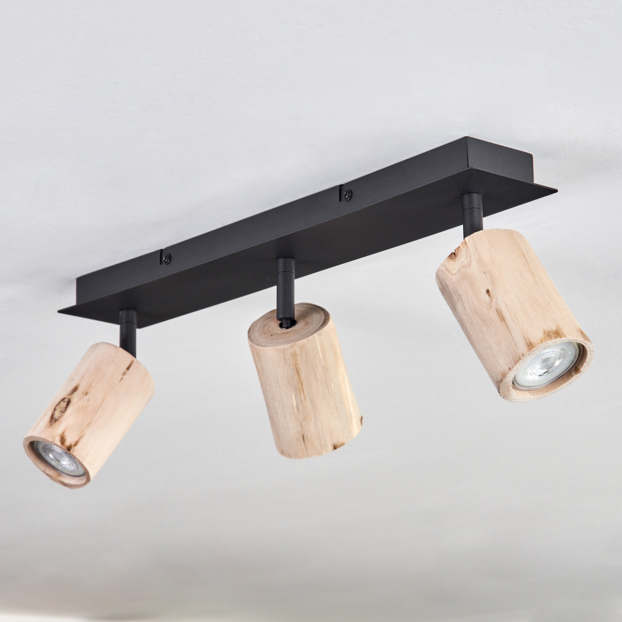 Eine Deckenleuchte mit drei zylindrischen Holz-Strahlern an einer schwarzen Metallbasis an einer weißen Decke.