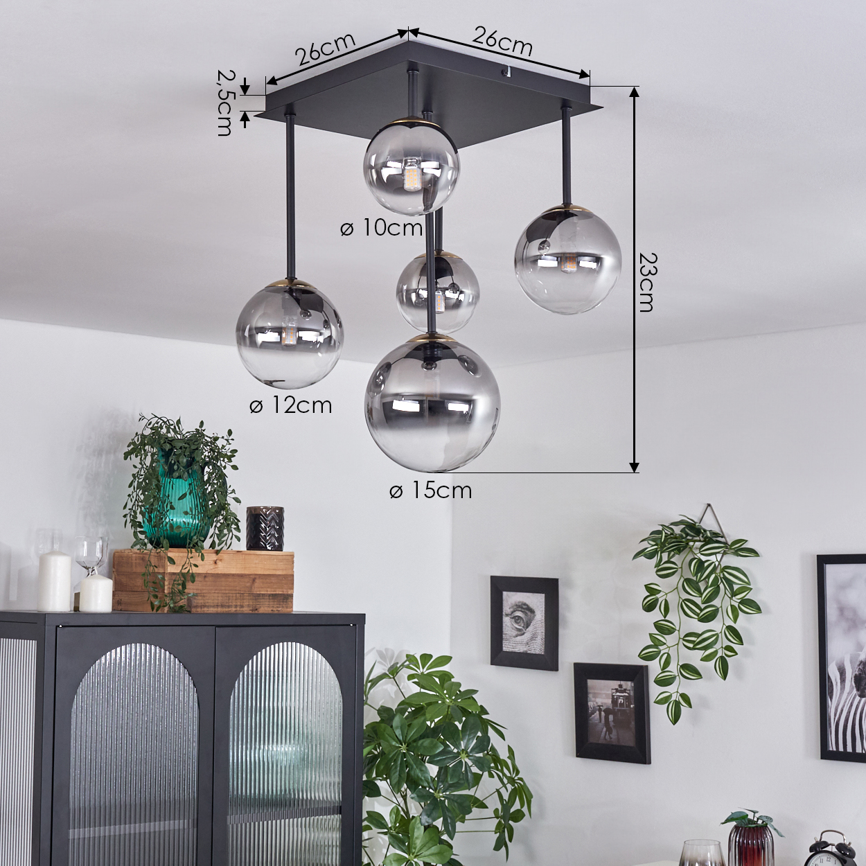 Eine schwarze Deckenlampe mit fünf unterschiedlich großen Glaskugeln. Maße sind angegeben. Pflanzen und Schrank im Hintergrund.
