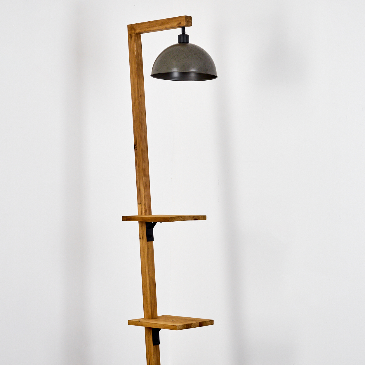 Eine Stehlampe aus Holz mit einem Lampenschirm aus Metall. Sie hat zwei Regale. Der Hintergrund ist eine weiße Wand.