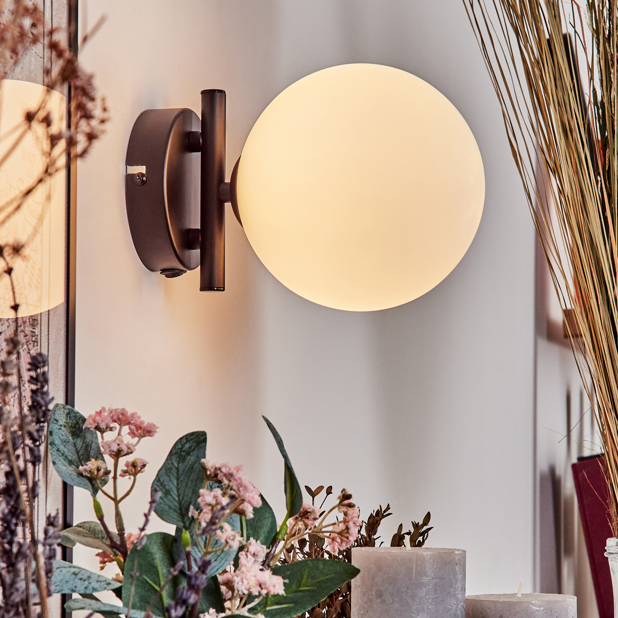 Eine Wandlampe mit rundem, leuchtendem Schirm. Blumen und Kerzen im Vordergrund. Trockenes Gras und ein Gemälde im Hintergrund.