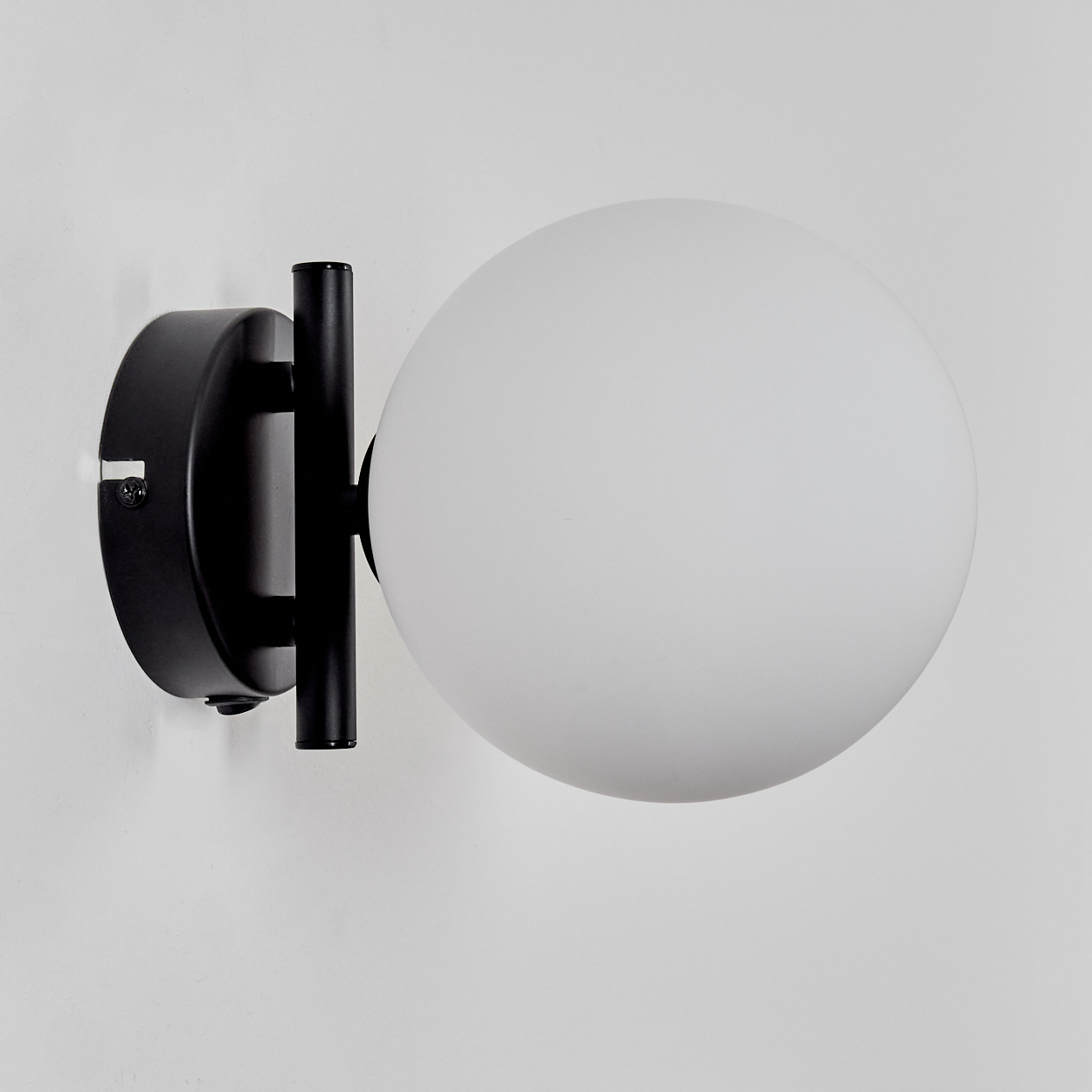 Eine Wandlampe mit schwarzer Metallstruktur und einem weißen, kugelförmigen Glasschirm vor einer weißen Wand.