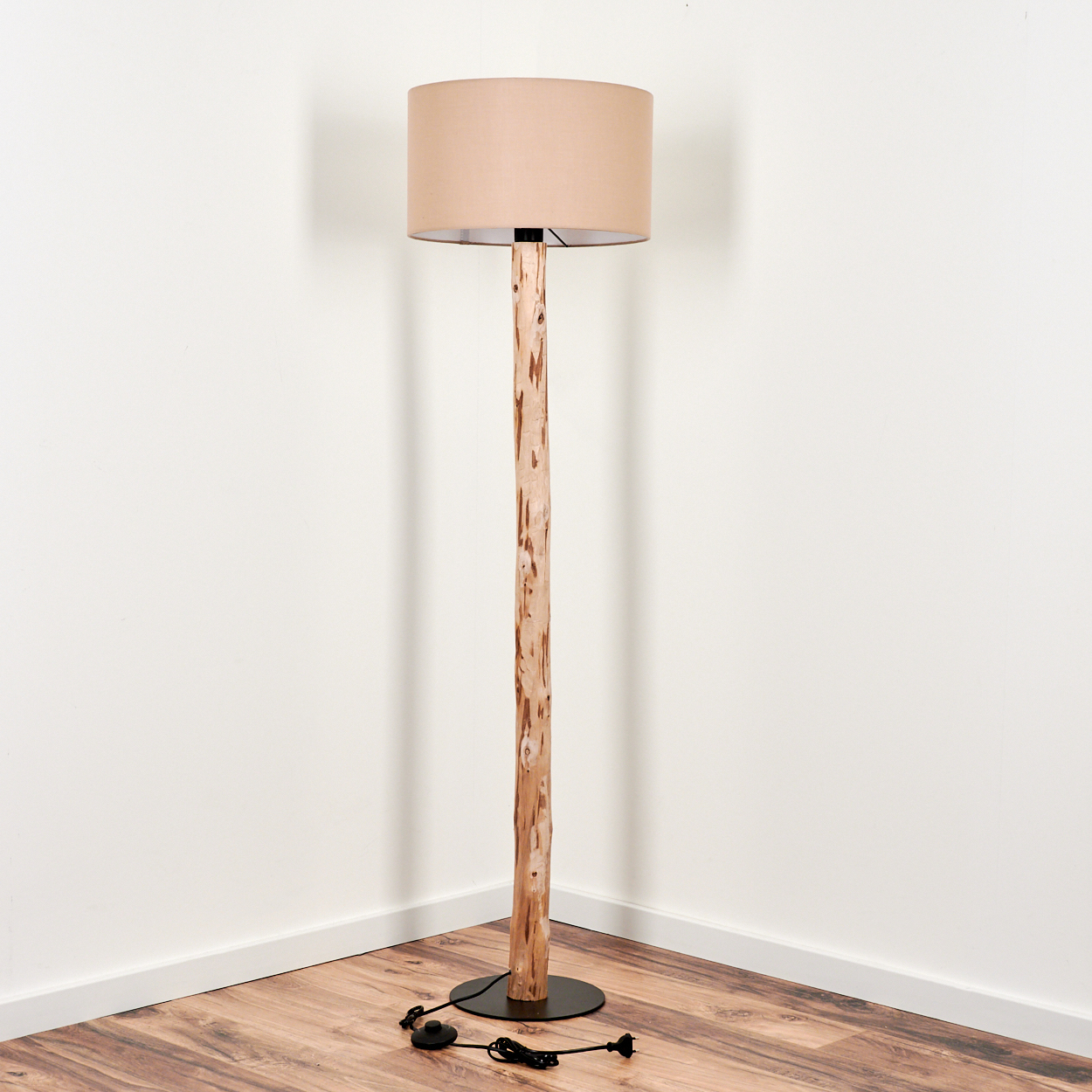 Eine Stehlampe mit beigem Lampenschirm und einem holzähnlichen Ständer. Sie steht auf einem schwarzen Sockel. Der Boden ist aus Holz, die Wände sind weiß.