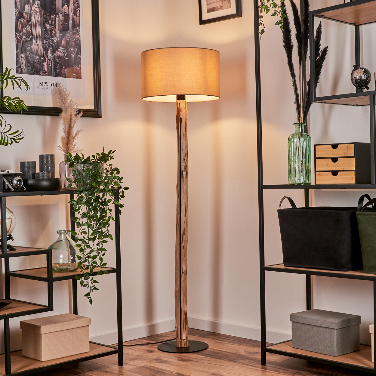 Eine hölzerne Stehlampe mit beigem Lampenschirm steht in einem Raum mit Regalen. Dekorative Gegenstände wie Pflanzen und Kerzen sind vorhanden.