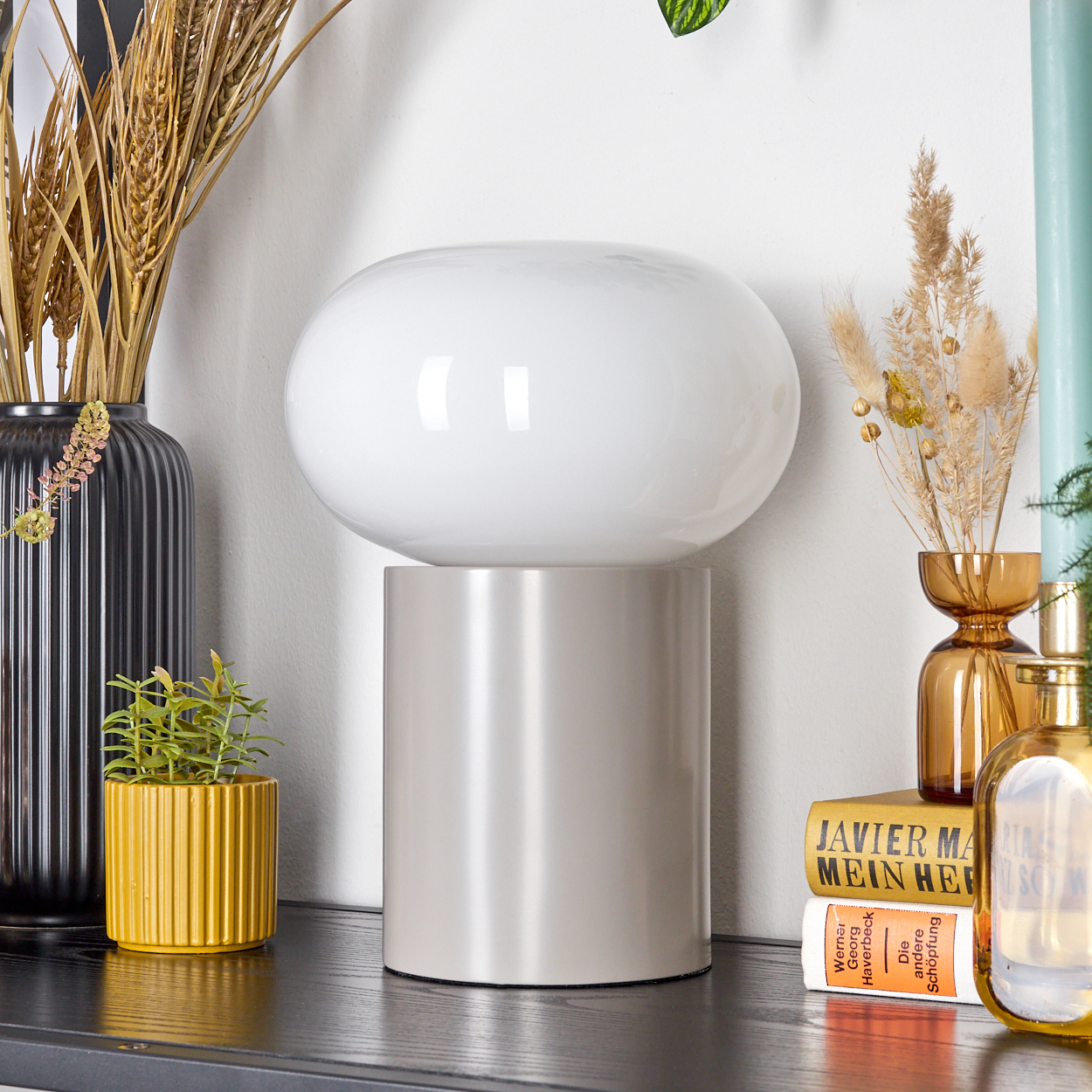 Eine weiße Lampe auf grauem Sockel. Es gibt einen gelben Blumentopf, eine schwarze Vase mit Weizen und dekorative Flaschen.