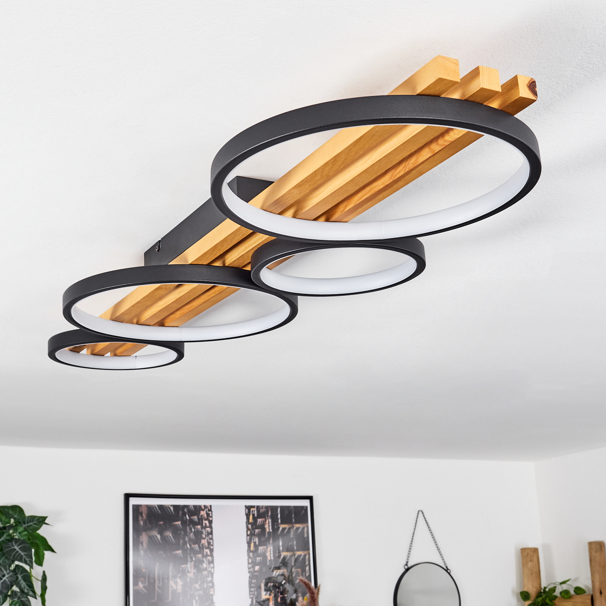 Eine moderne Deckenleuchte mit vier schwarzen, kreisförmigen LED-Leuchten, Holzbalken und einer weißen Decke. Ein gerahmtes Bild hängt unten.