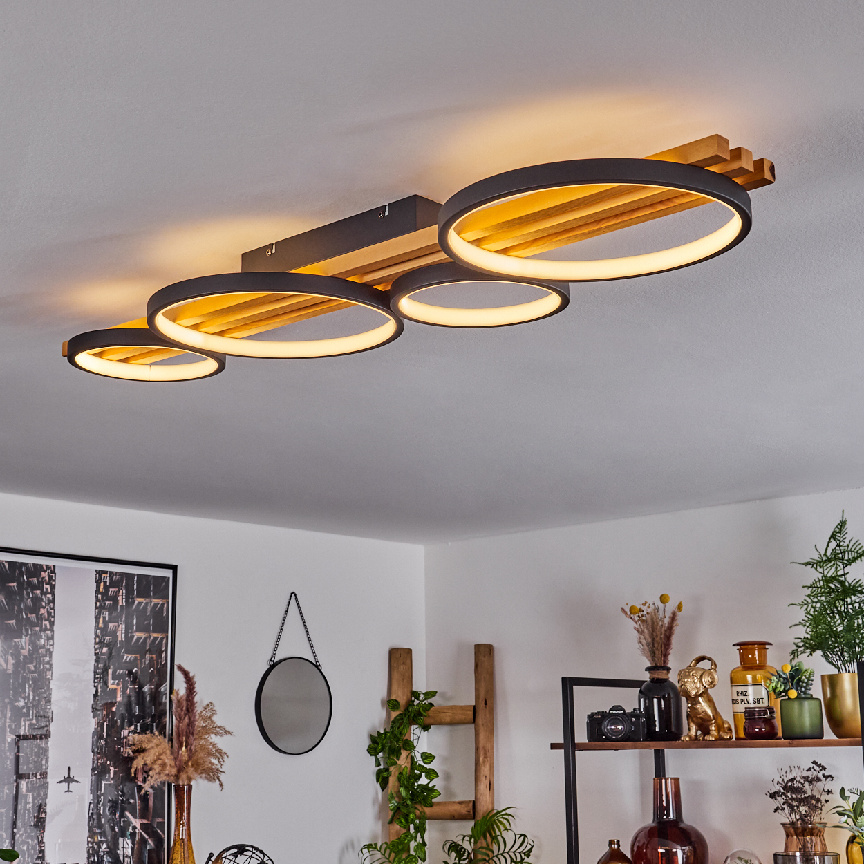Moderne Deckenleuchte mit vier beleuchteten Ringen, Holzakzenten. Raum mit Deko, Spiegel, Pflanzen und Regalen mit verschiedenen Gegenständen.
