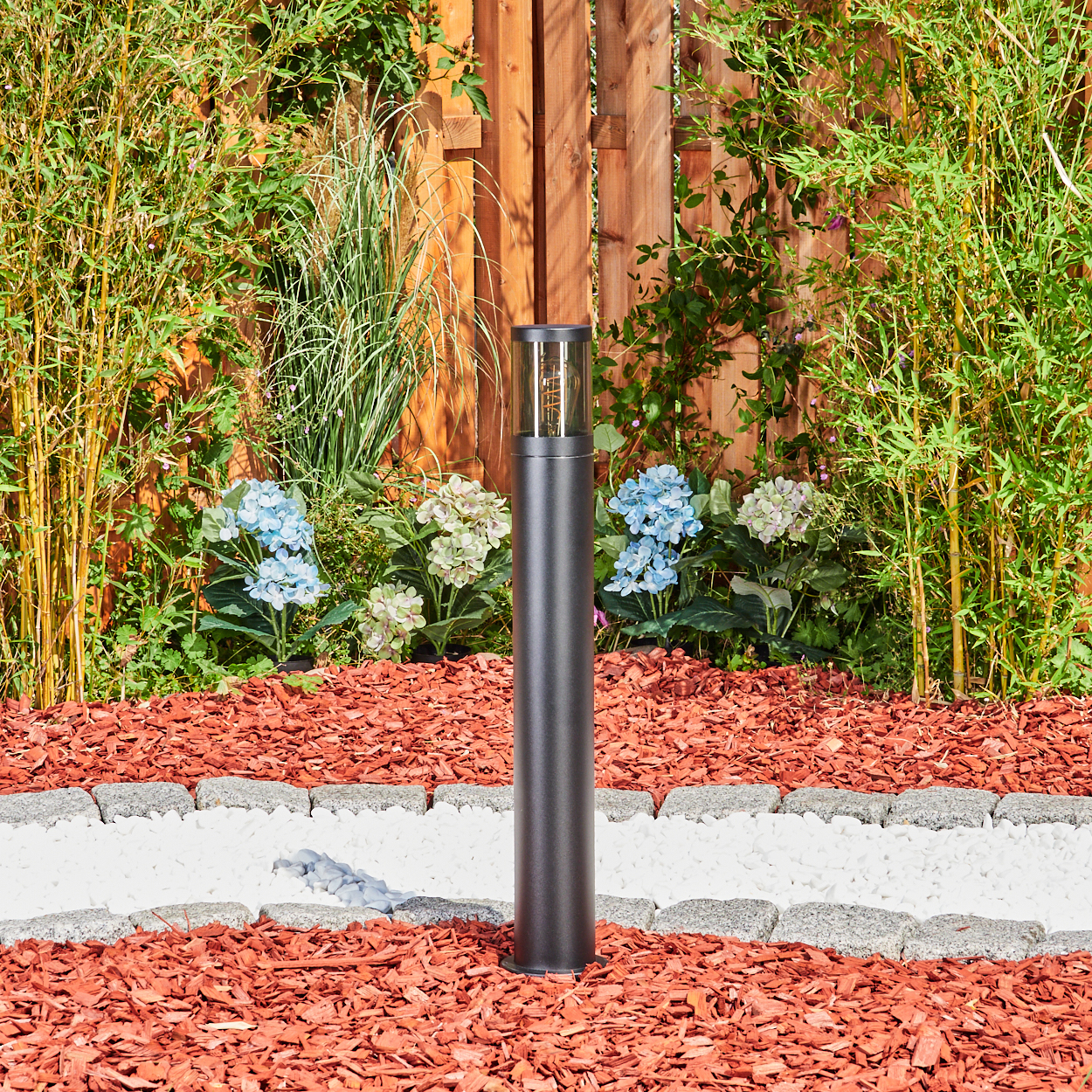 Eine schwarze Außenlampe steht in einem Gartenbeet. Das Beet hat roten Mulch, begrenzt von Steinen und weißem Kies. Grüne Pflanzen umgeben.