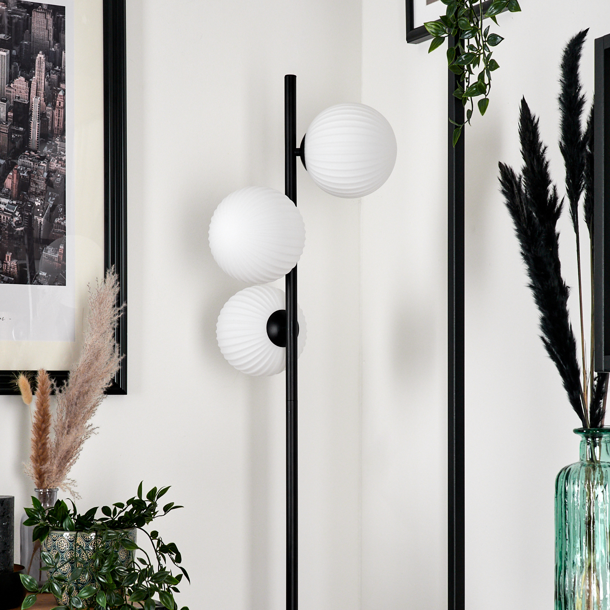 Eine moderne schwarze Stehlampe mit drei weißen Kugelschirmen. Dekorative Pflanzen in Vasen stehen daneben, mit einem Stadtbilddruck im Hintergrund.