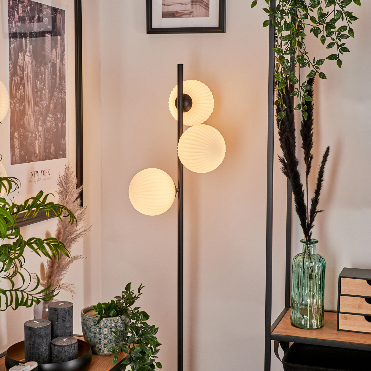 Ein modernes Zimmer mit Stehlampe. Es hat drei kugelförmige Lampen. Es gibt Pflanzen, Kerzen und ein gerahmtes Bild.