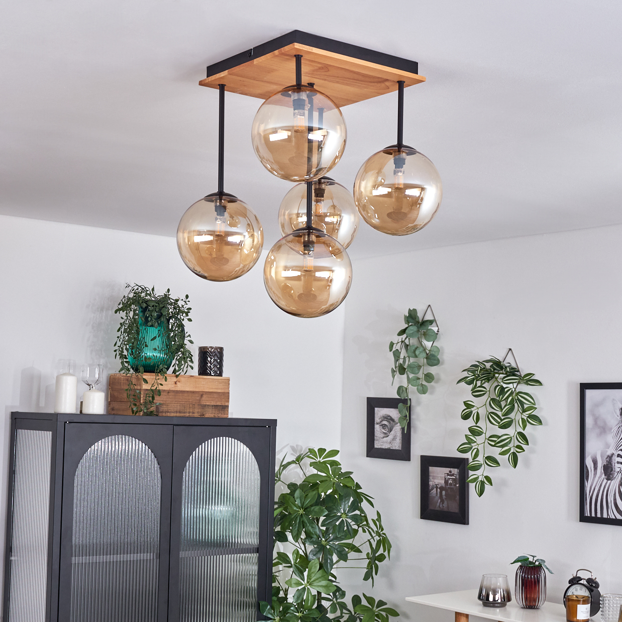 Eine moderne Leuchte mit fünf bernsteinfarbenen Glaskugeln hängt an einer Holzdecke. Dekorative Pflanzen und Möbel umgeben das Ganze.