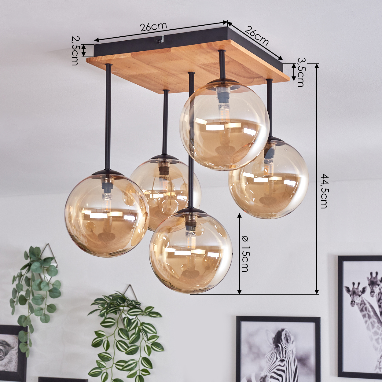 Eine Deckenlampe mit fünf bernsteinfarbenen Glaskugeln, schwarzen Stäben und einem Holzsockel. Maße sind angegeben. Grüne Pflanzen und gerahmte Kunst sind sichtbar.