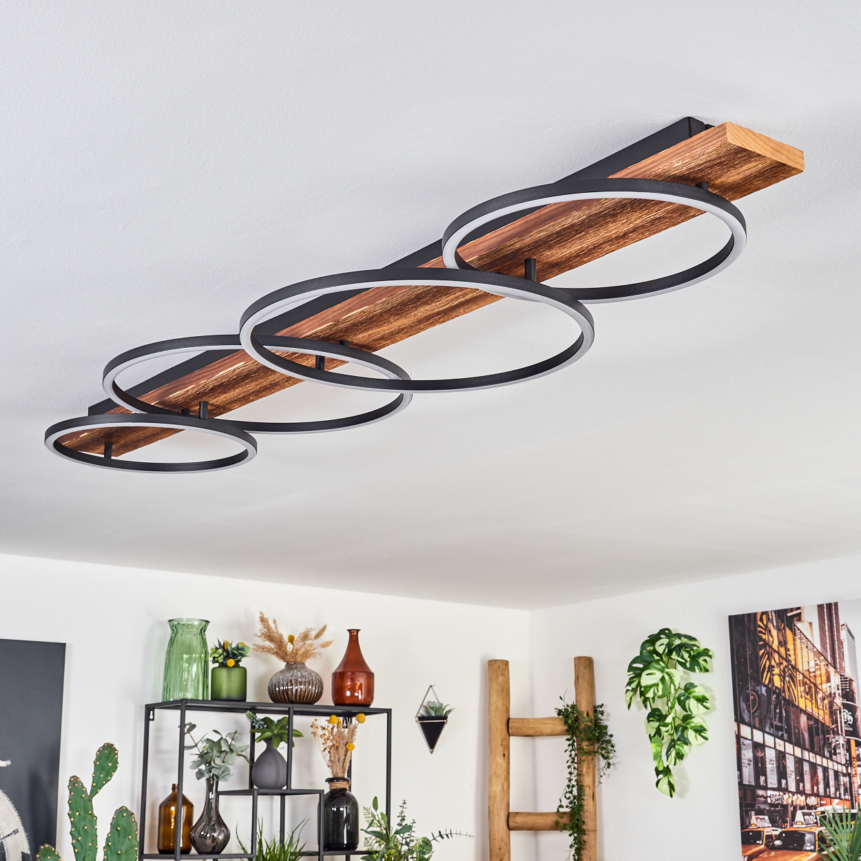 Eine moderne Deckenleuchte mit vier schwarzen, kreisförmigen Lichtern, die auf einer Holzplanke montiert sind. Darunter sind Regale mit Vasen und Pflanzen zu sehen.