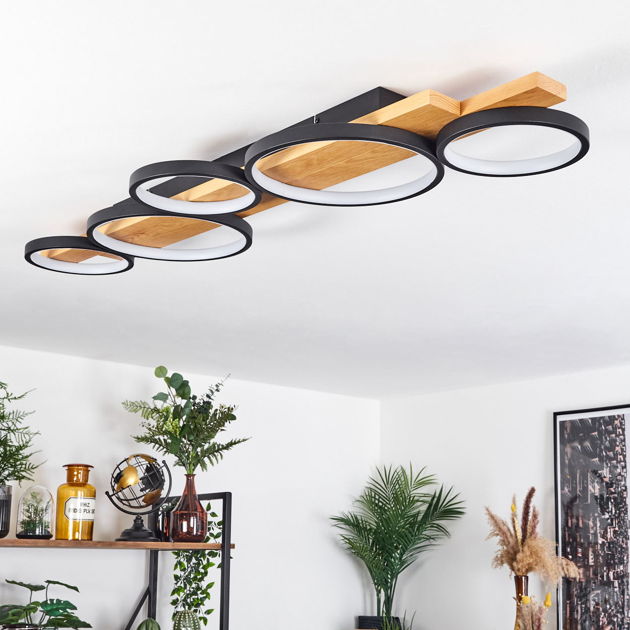 Moderne Deckenleuchte mit schwarzen Ringen und Holzbalken. Pflanzen und Dekoration sind unten vor einer weißen Wand zu sehen.