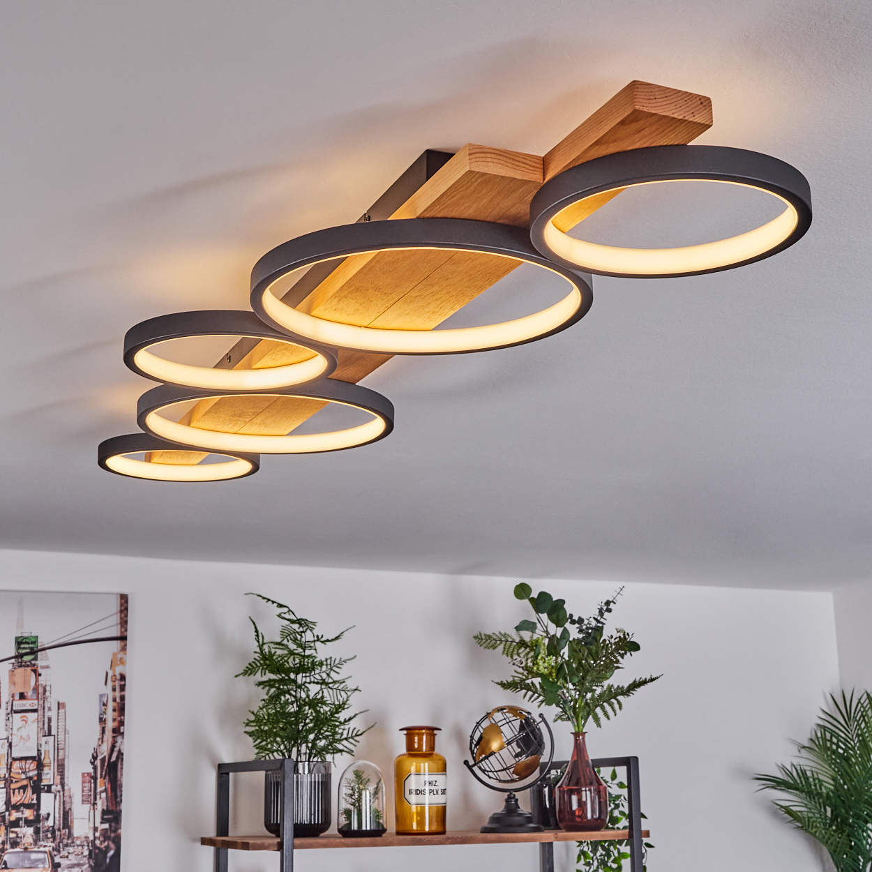 Moderne Deckenleuchte mit fünf beleuchteten Ringen. Holzbalken tragen die Leuchte. Regale unten zeigen Pflanzen, Globus und Dekoration.