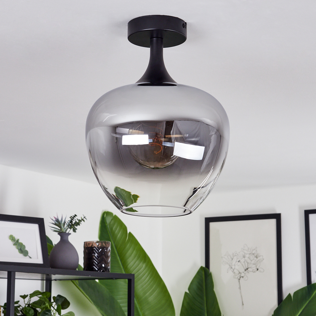 Eine moderne, graue Glas-Hängeleuchte hängt von einer weißen Decke. Darunter dekorieren Pflanzen und gerahmte Kunst den Raum.