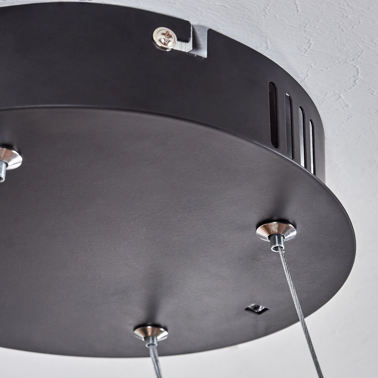 Nahaufnahme einer schwarzen Deckenleuchte. Silberne Kabel und Schrauben sind sichtbar. Der Hintergrund ist eine strukturierte, hellfarbige Decke.