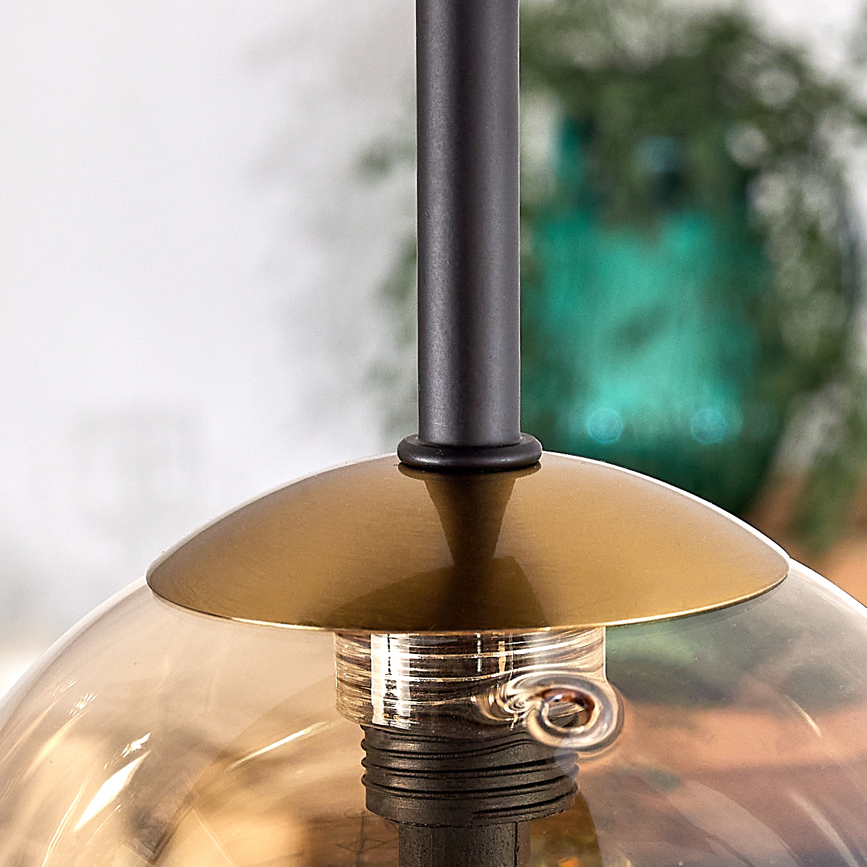 Nahaufnahme einer Hängeleuchte. Sie hat eine schwarze Stange, einen goldenen Schirm und eine klare, bernsteinfarbene Glaskugel. Der Hintergrund ist unscharf.