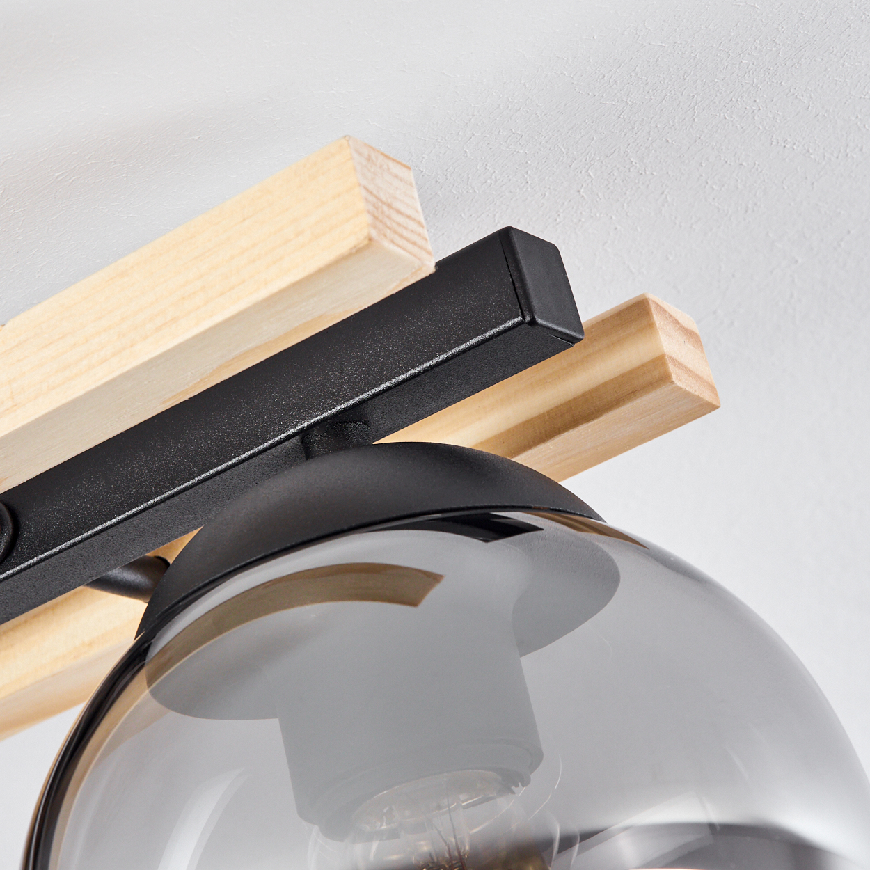 Nahaufnahme einer Lampe. Sie hat einen Glasschirm, einen schwarzen Metallrahmen und Holzbalken. Der Hintergrund ist eine schlichte weiße Decke.