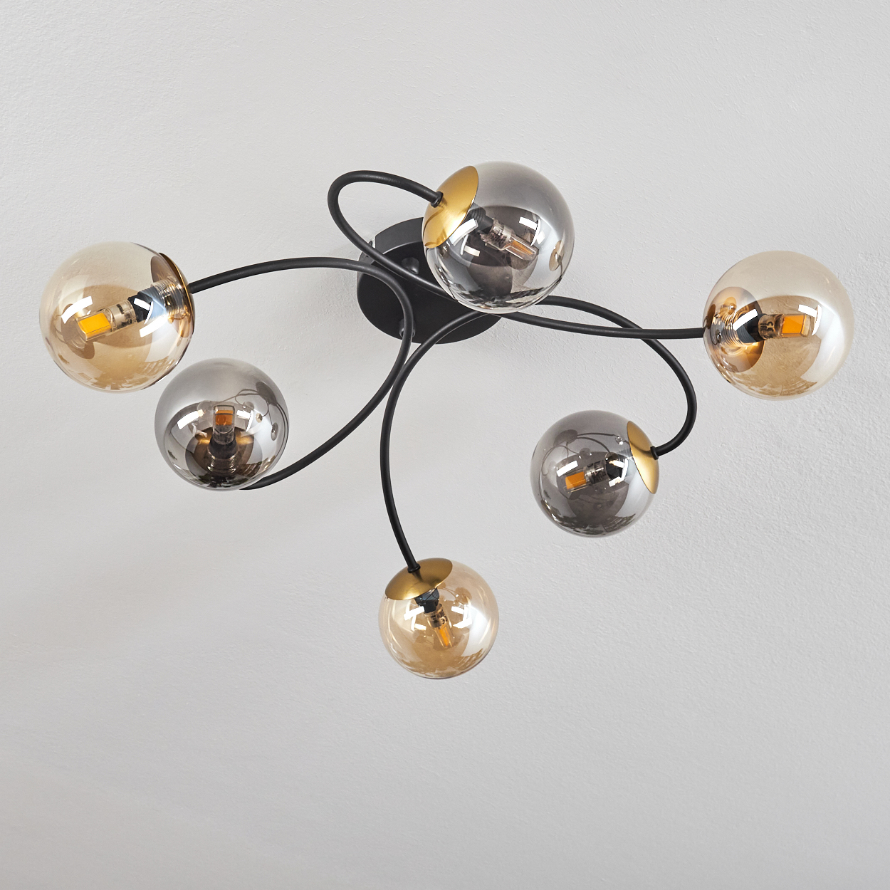Eine Deckenlampe mit schwarzen, gebogenen Armen und sechs kugelförmigen Lampenschirmen in Gold- und Silberfarben vor weißer Decke.