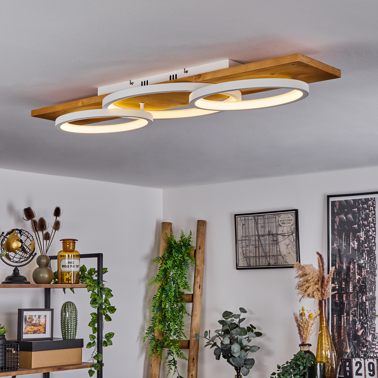 Eine moderne Deckenlampe mit drei kreisförmigen Lichtern ist auf einem Holzbrett montiert. Darunter ist ein Raum mit Pflanzen und Kunst dekoriert.