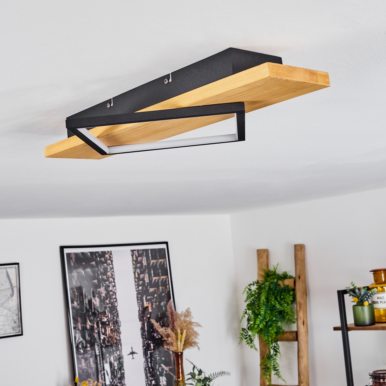 Eine moderne Deckenlampe mit Holzbalken und schwarzem Metallrahmen. Darunter ein Raum mit Kunstwerken, Pflanzen und einem leiterartigen Regal.