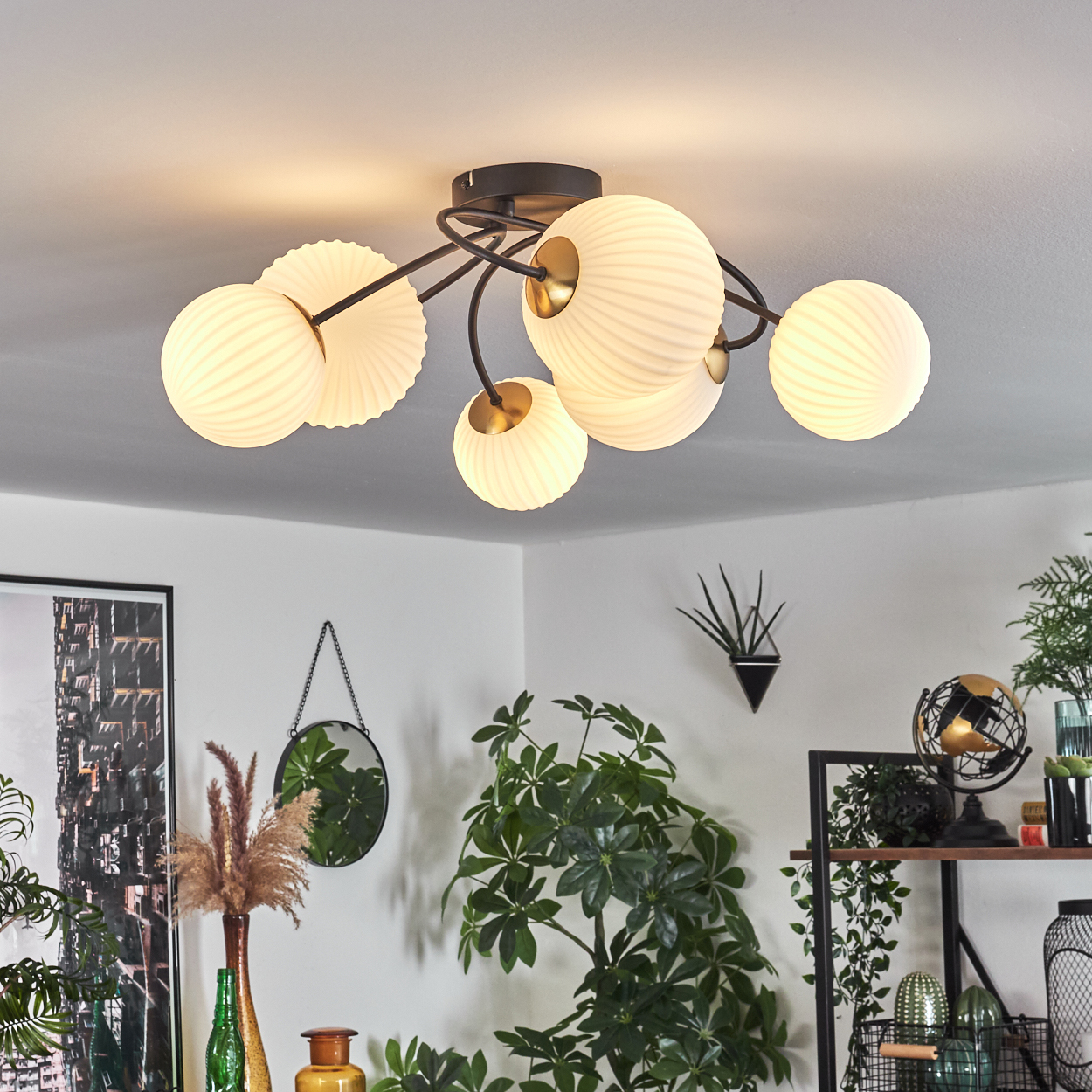 Deckenlampe mit weißen Kugeln und dunklem Rahmen. Pflanzen, Spiegel und Kunstwerk im Raum. Die Szene ist gut beleuchtet.