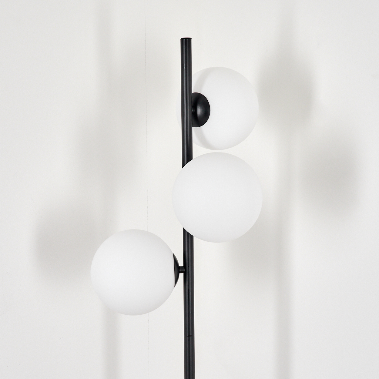 Eine moderne schwarze Stehlampe mit drei weißen Kugelleuchten. Die Lampe steht vor einer schlichten weißen Wand und wirft einen Schatten.