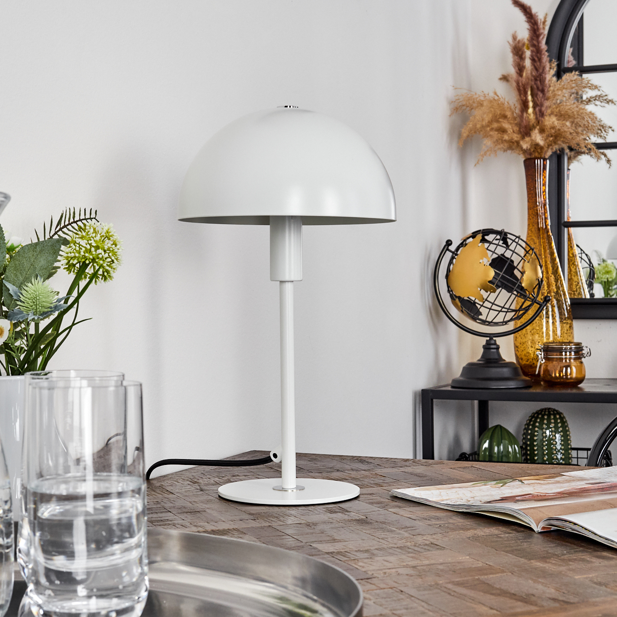 Eine weiße Lampe auf einem Holztisch, mit Wassergläsern, Blumen und einem Globus im Hintergrund.