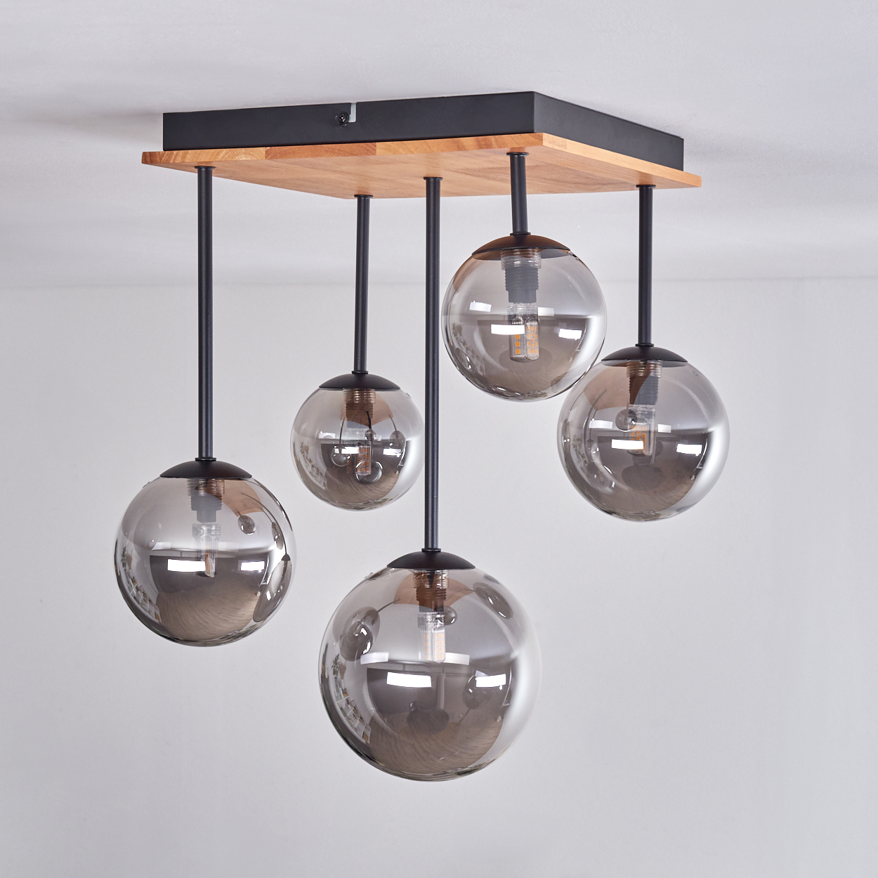 Eine moderne Deckenleuchte mit fünf rauchigen Glaskugelschirmen, die an einer Holz- und schwarzen Metallvorrichtung hängen. Weisse Wand im Hintergrund.