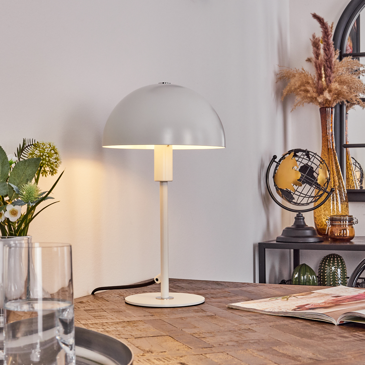 Graue Lampe auf einem Holztisch. Eine Vase mit Blumen, ein Globus und eine Zeitschrift sind ebenfalls auf dem Tisch. Weiches Licht.