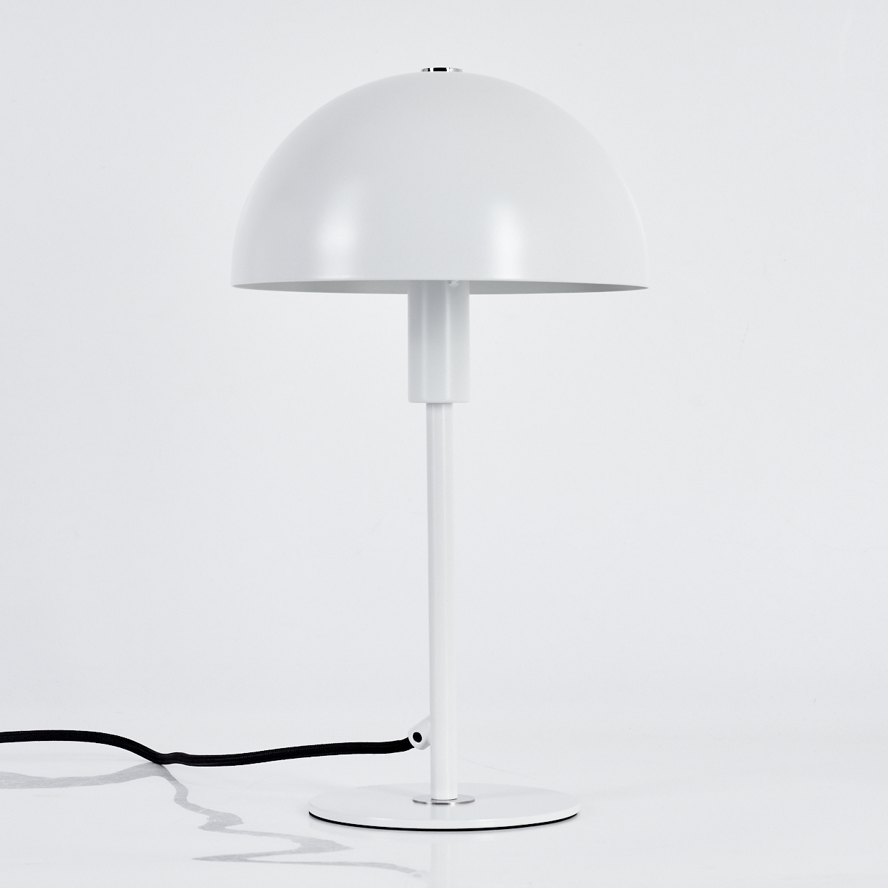 Eine weiße Schreibtischlampe mit geschwungenem Lampenschirm, dünnem Stiel und kreisförmigem Sockel. Ein schwarzes Netzkabel geht von der Basis aus.