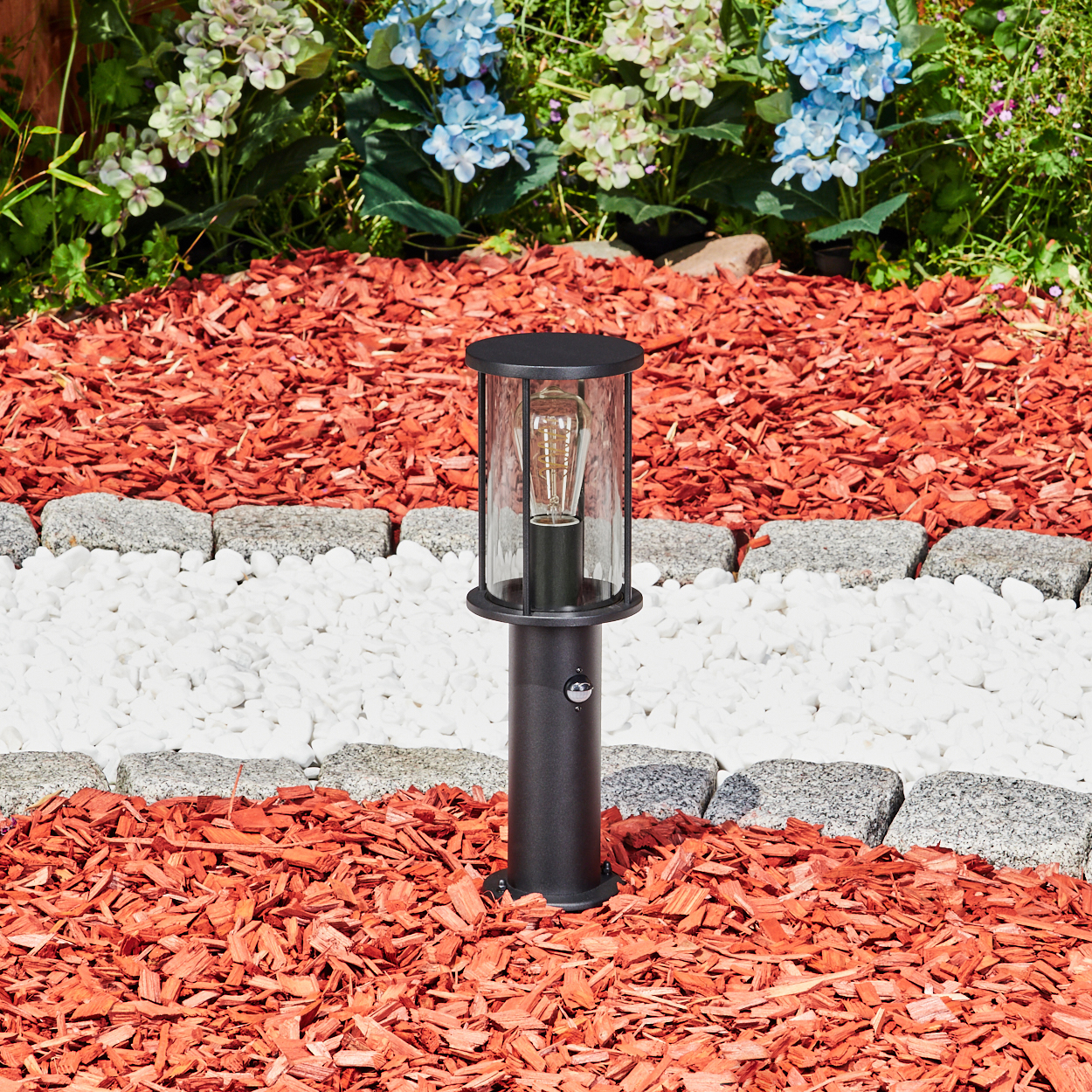 Eine schwarze Außenlampe mit Glasgehäuse steht auf roter Mulch. Weiße Steine und graue Steine bilden eine Grenze. Im Hintergrund blühen Blumen.