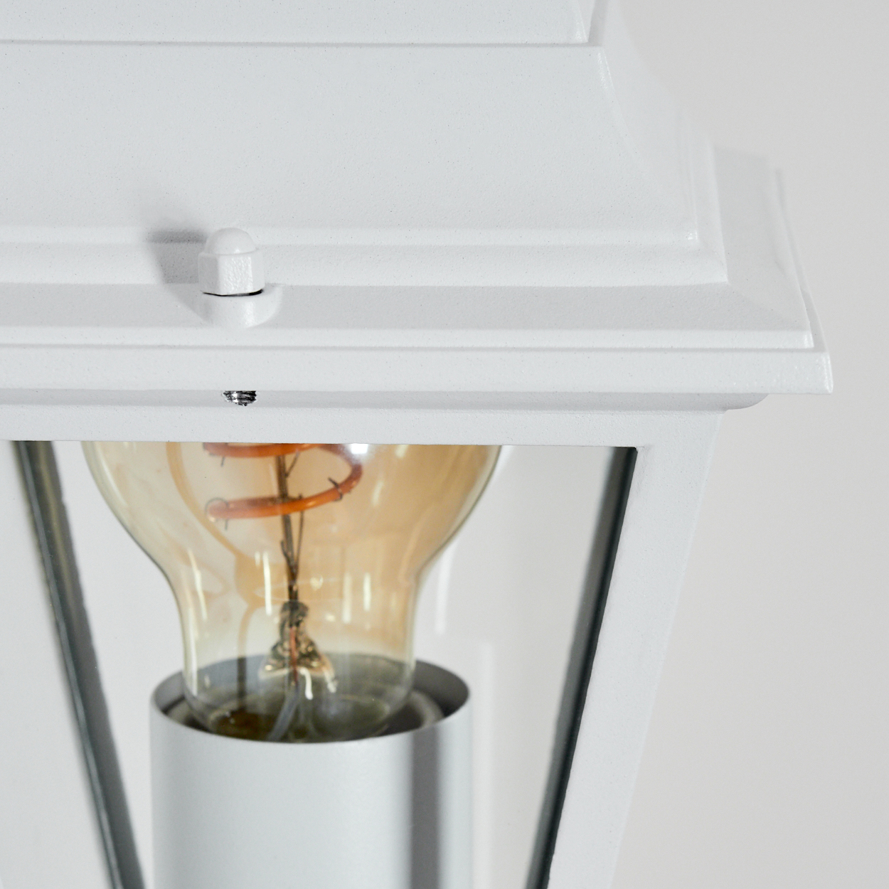 Nahaufnahme einer weißen Außenlampe. Im Inneren ist eine Glühbirne mit orangefarbenem Glühfaden sichtbar. Der Hintergrund ist eine schlichte, weiße Wand.