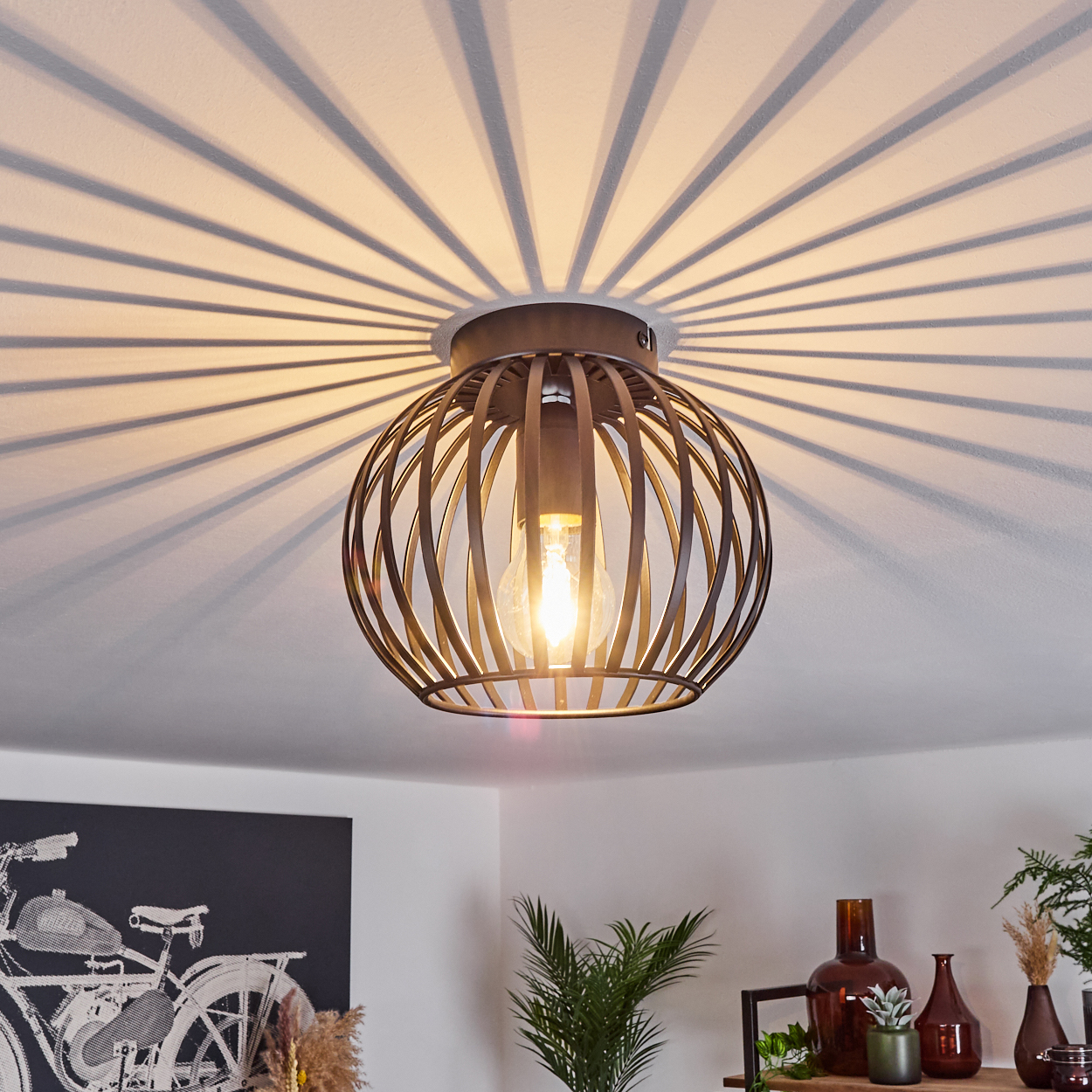 Eine kugelförmige Metallkäfig-Leuchte mit einer brennenden Glühbirne hängt von der Decke und wirft radiale Schatten. Ein Fahrrad-Kunstwerk ist sichtbar.