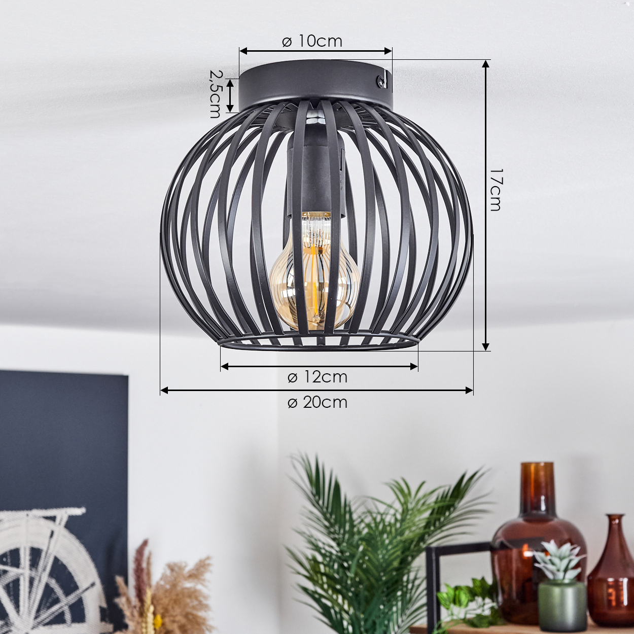 Schwarze, käfigartige Deckenlampe mit sichtbarer Glühbirne. Maße sind angegeben. Pflanzen und Vasen im Hintergrund.