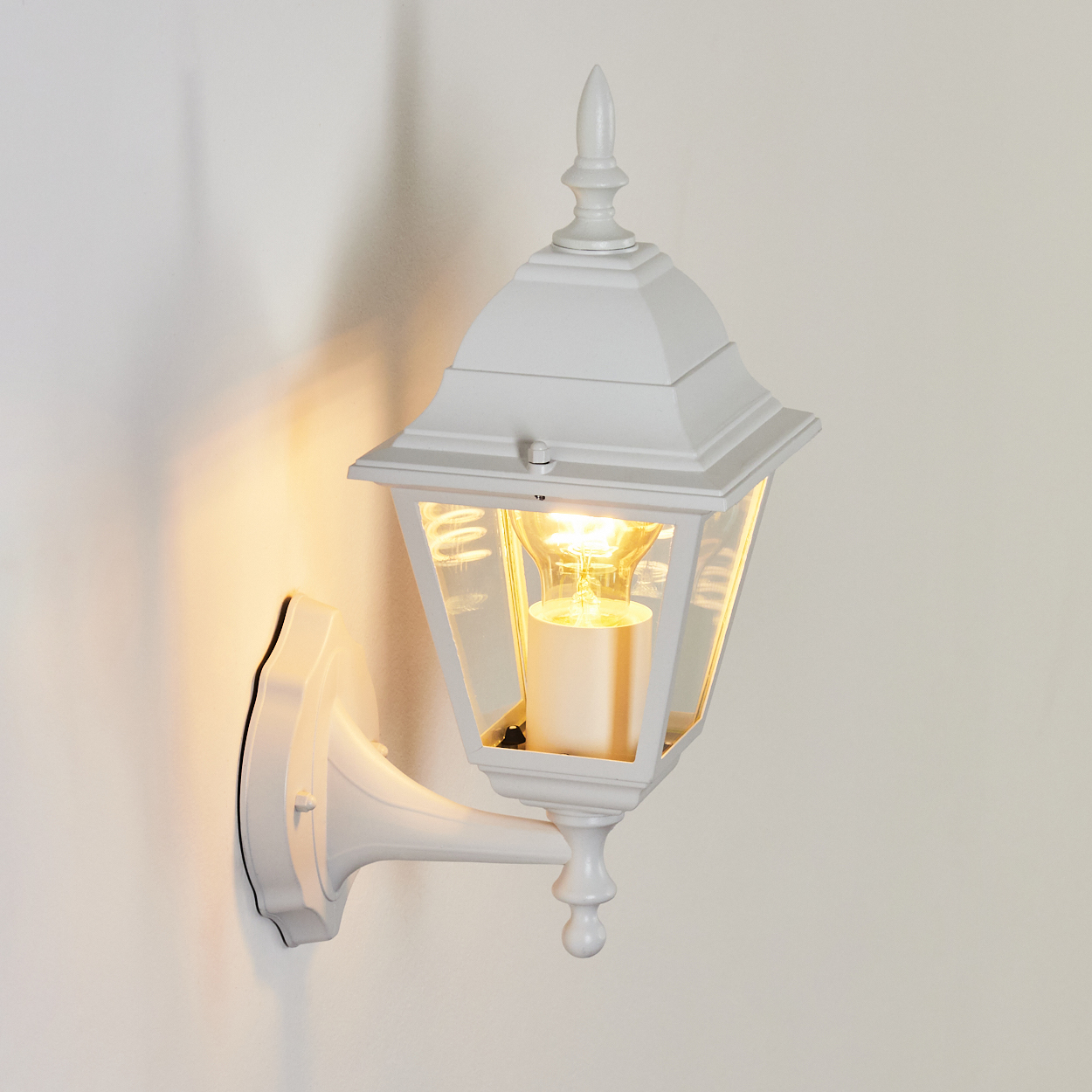 Eine weiße Außenlaterne mit einer brennenden Glühbirne ist an einer weißen Wand montiert. Die Lampe hat ein klassisches Design und klares Glas.