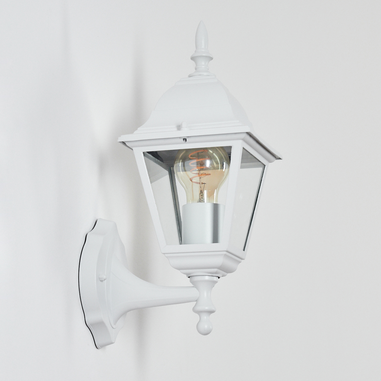 Eine weiße Wandleuchte mit Glasgehäuse und Glühbirne. Es ist an einer weißen Wand montiert.