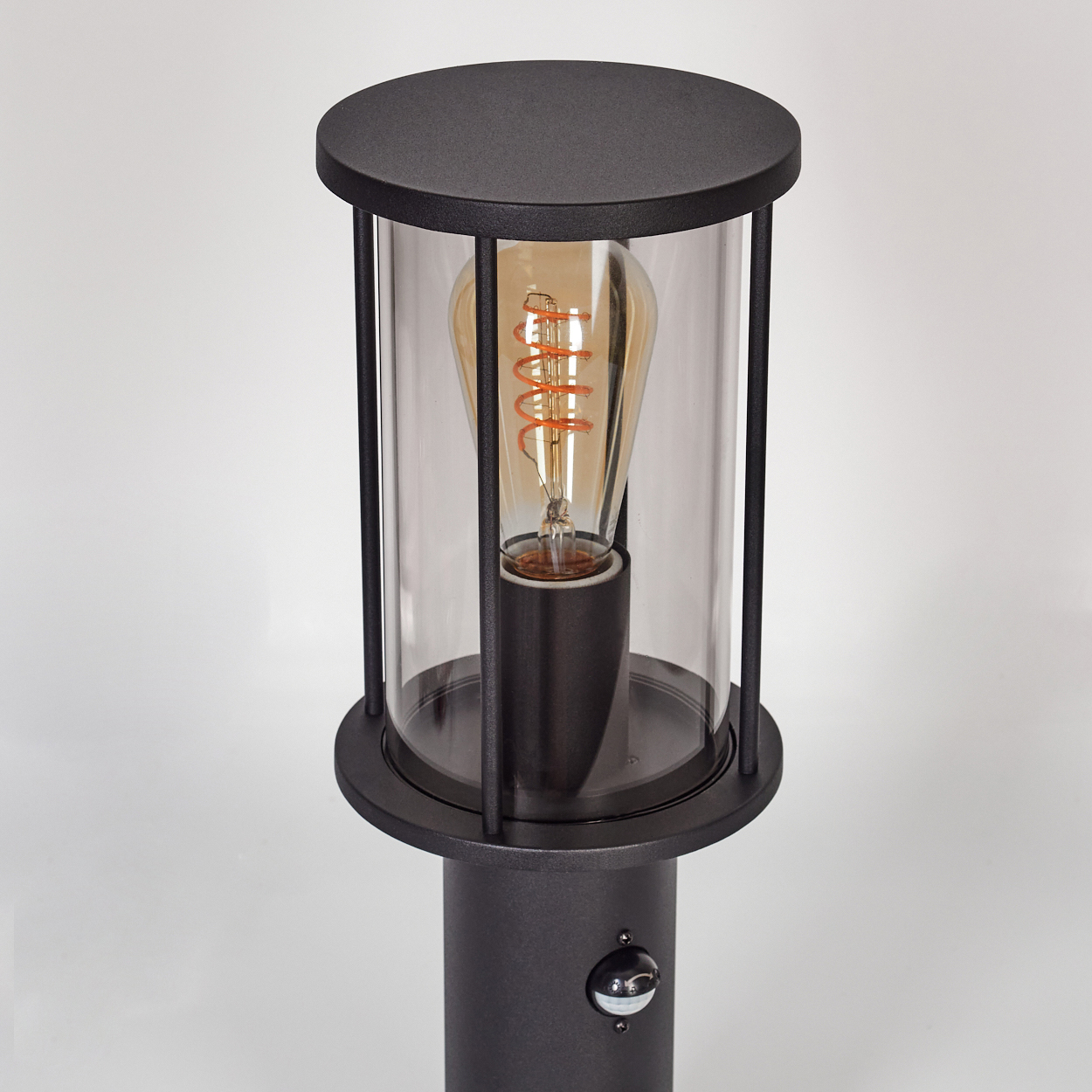 Eine schwarze Außenleuchte mit einem durchsichtigen Glasgehäuse und einer dekorativen Glühbirne im Inneren. Die Lampe steht auf einem grauen Pfahl, mit einem Sensor.