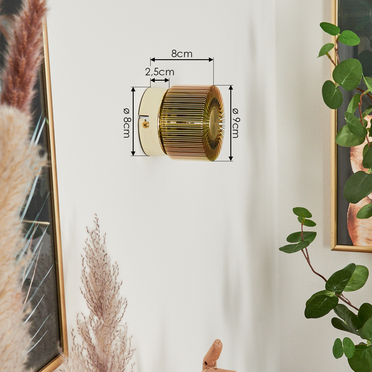 Eine zylindrische Wandleuchte mit goldenen Lamellen, Maße markiert. Daneben ein gerahmtes Kunstwerk und eine Pflanze mit grünen Blättern.